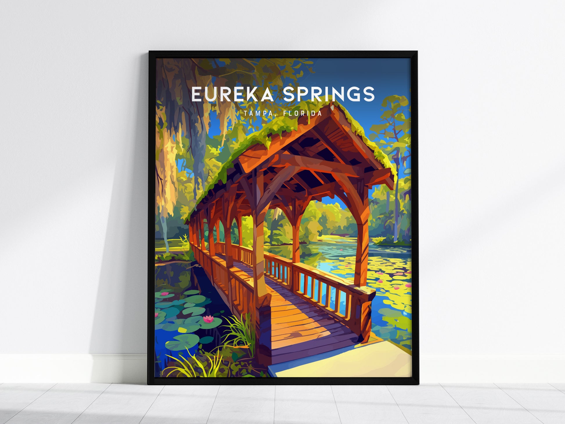 A framed poster of Eureka Springs, Tampa, depicts a covered wooden bridge over a tranquil stream surrounded by lush greenery and water lilies under a clear blue sky.