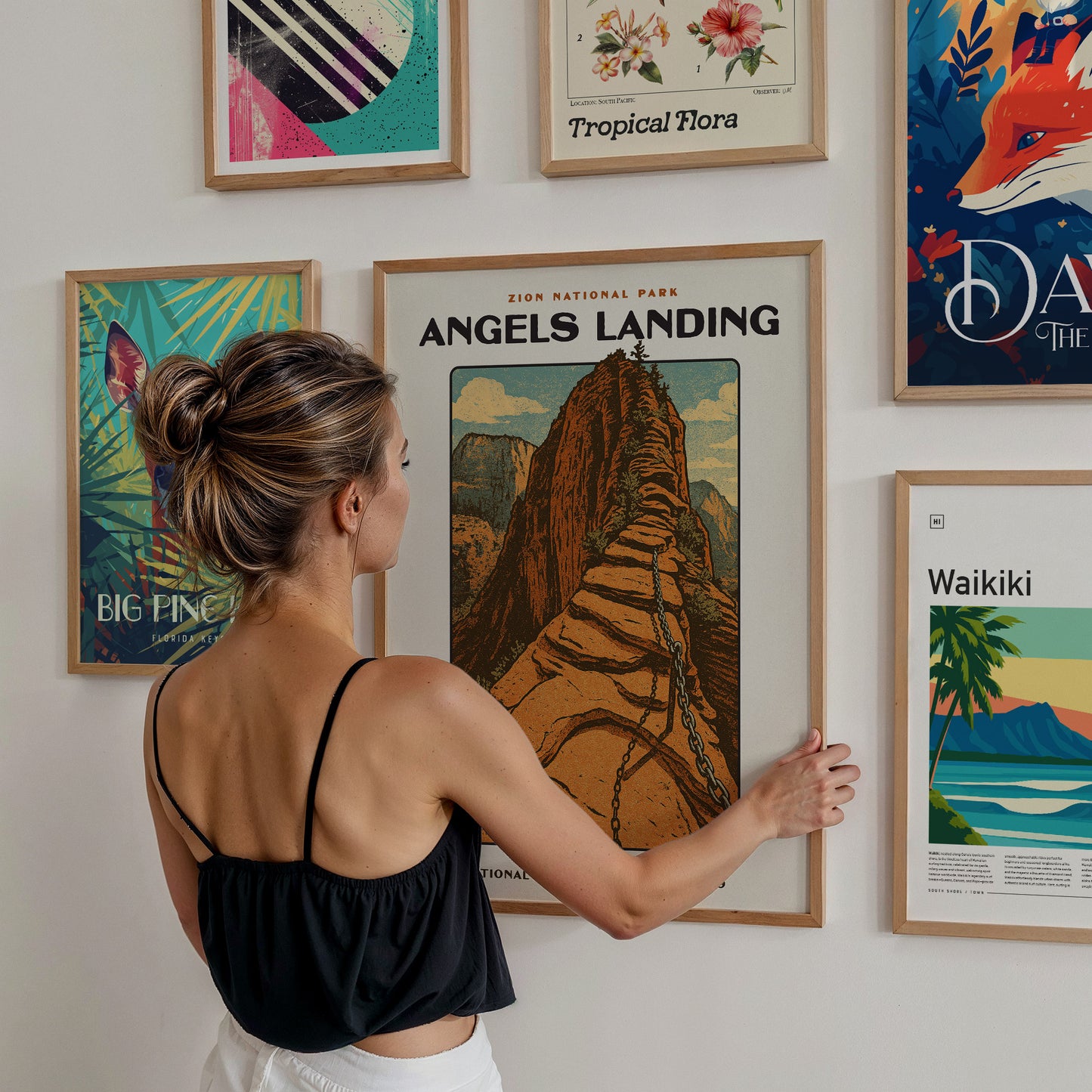 A woman with long hair tied in a bun is admiring a framed poster of Angels Landing in Zion National Park, which features a stylized illustration of a rocky mountain path.