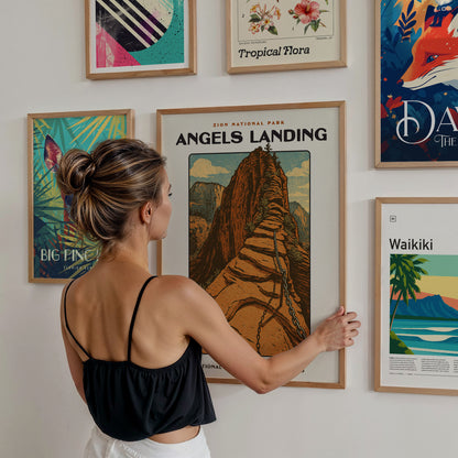 A woman with long hair tied in a bun is admiring a framed poster of Angels Landing in Zion National Park, which features a stylized illustration of a rocky mountain path.