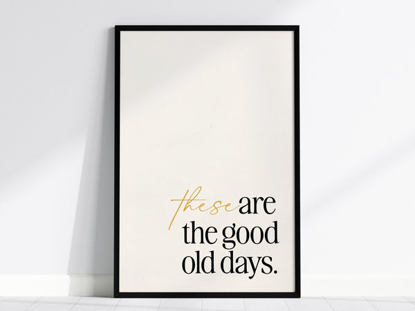 A minimalist poster with the phrase these are the good old days in elegant typography, featuring a mix of gold and black text. The artwork is framed in black and rests against a white wall on a light floor.