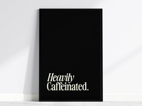 A minimalist poster, wall art featuring the phrase Heavily Caffeinated in white typography on a black background. The artwork is leaning against a white wall, creating a striking contrast.