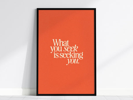 A framed poster with the quote What you seek is seeking you. is displayed against a white wall. The text is in bold, white typography on a vibrant orange background, creating a striking contrast.