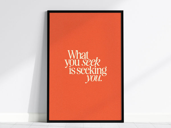 A framed poster with the quote What you seek is seeking you. is displayed against a white wall. The text is in bold, white typography on a vibrant orange background, creating a striking contrast.