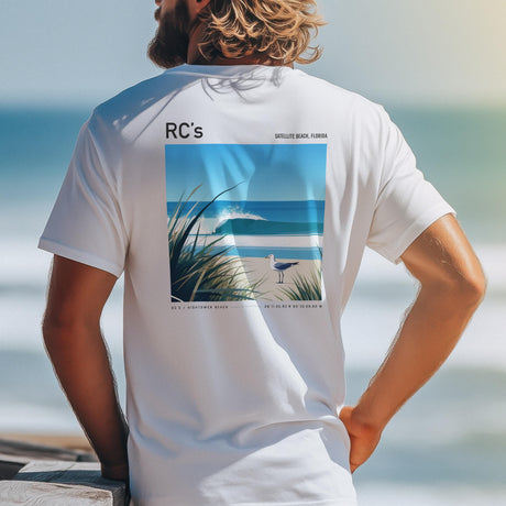 A man with long hair is wearing a white shirt featuring a poster-style beach scene with waves and a seagull, labeled RCs Satellite Beach, Florida. The shirt acts as wall art, capturing the essence of a coastal vibe.