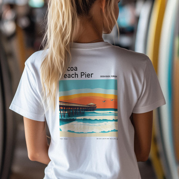 A person with long blonde hair is wearing a white T-shirt featuring a colorful poster-style graphic of Cocoa Beach Pier at sunset. The image highlights waves and birds, capturing the essence of Florida wall art.
