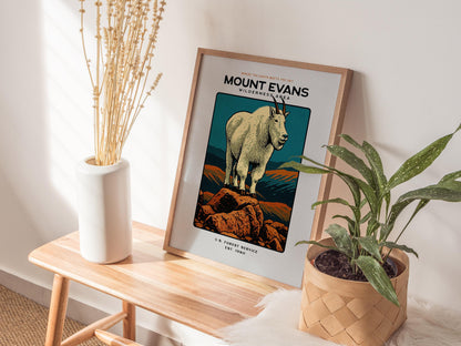 Framed poster of a mountain goat on Mount Evans with plants and a vase on a wooden table.