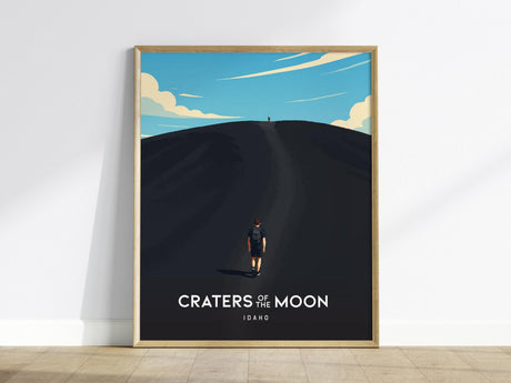 This poster, featuring wall art of the Craters of the Moon in Idaho, shows a lone hiker climbing a black cinder cone under a vast blue sky with clouds. The artwork is styled in a vintage-modern design within a wooden frame.