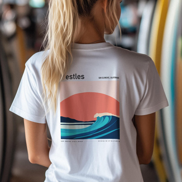 A person with long blonde hair is wearing a white t-shirt featuring a colorful surf art design on the back. The artwork depicts a wave against a sunset backdrop, labeled with San Clemente, California. Ideal for surf-themed poster, wall art.