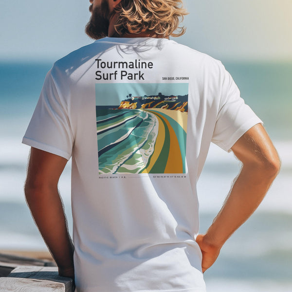 A man with long hair stands by the ocean, wearing a white t-shirt featuring a colorful Tourmaline Surf Park design. The shirt showcases vibrant waves and cliffs, resembling poster wall art.