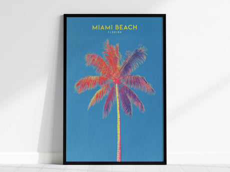 A vibrant poster features a colorful, stylized palm tree against a bright blue background, with the words Miami Beach Florida in yellow at the top. The artwork is framed in black and leans against a white wall.