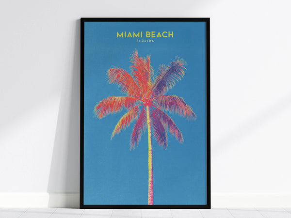 A vibrant poster features a colorful, stylized palm tree against a bright blue background, with the words Miami Beach Florida in yellow at the top. The artwork is framed in black and leans against a white wall.