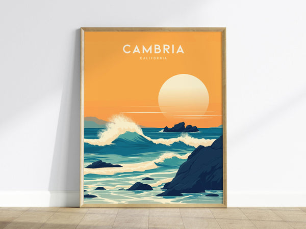 A framed poster of Cambria, California showcases a stylized coastal sunset. The artwork features rolling waves crashing against rugged rocks under a large, setting sun, with an orange and blue color palette.