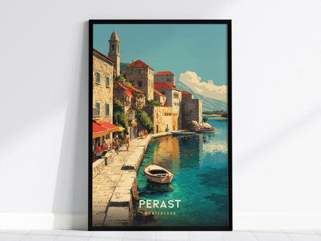 The image shows a framed poster depicting a coastal scene of Perast, Montenegro. It features stone buildings with red roofs along a waterfront, a small boat on the clear blue water, and a mountainous backdrop under a clear sky.