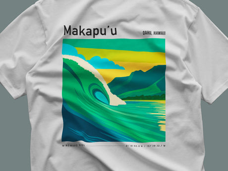 The image shows a white t-shirt featuring a vibrant graphic of a large wave with lush green mountains and a bright sky in the background. The text Makapuu Oahu, Hawaii is printed above the graphic.