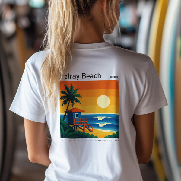 A person with long blonde hair is wearing a white T-shirt featuring a colorful graphic on the back. The design includes a sunset, waves, a palm tree, and a lifeguard tower, with the text Delray Beach Florida.
