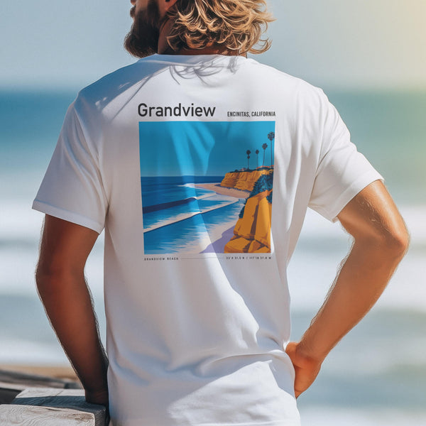 A person with wavy hair stands by the ocean, wearing a white t-shirt featuring a colorful illustration of Grandview Beach with cliffs and palm trees. The shirt reads Grandview, Encinitas, California.