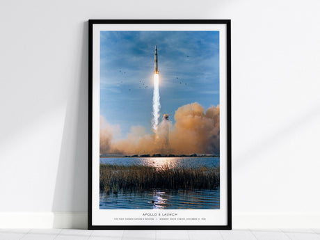 A framed print depicts the Apollo 8 launch with the Saturn V rocket ascending against a blue sky, leaving a trail of smoke. The scene includes birds flying near the rocket and reflections on the water below.