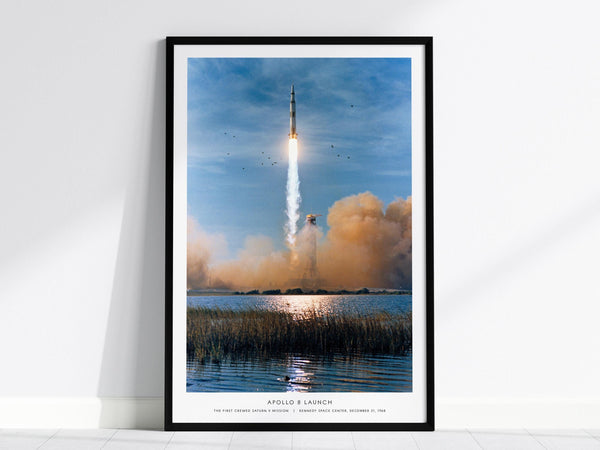 A framed print depicts the Apollo 8 launch with the Saturn V rocket ascending against a blue sky, leaving a trail of smoke. The scene includes birds flying near the rocket and reflections on the water below.