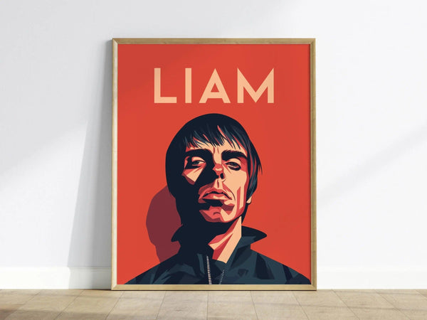 A framed poster features a stylized portrait of a person against a bold red background with LIAM written above. The artwork is propped against a white wall on a wooden.