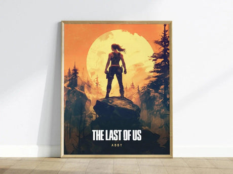 A framed poster featuring a silhouetted figure of Abby standing on a rock against a vibrant orange sunset, surrounded by dark trees. The text below reads THE LAST OF US and ABBY, capturing the games intense and emotional themes.