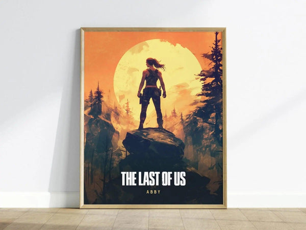 A framed poster featuring a silhouetted figure of Abby standing on a rock against a vibrant orange sunset, surrounded by dark trees. The text below reads THE LAST OF US and ABBY, capturing the games intense and emotional themes.