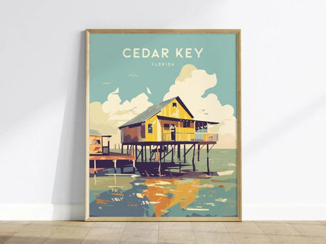 A vibrant art print of Cedar Key, Florida, featuring a classic stilt house above calm waters, framed in light wood. The illustration showcases soft coastal tones and stylized clouds, evoking the charm of Old Florida.