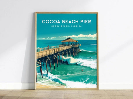 A framed poster depicts Cocoa Beach Pier in Florida, extending over the ocean with a thatched-roof pavilion and surfers riding waves underneath. The sky is clear and blue.