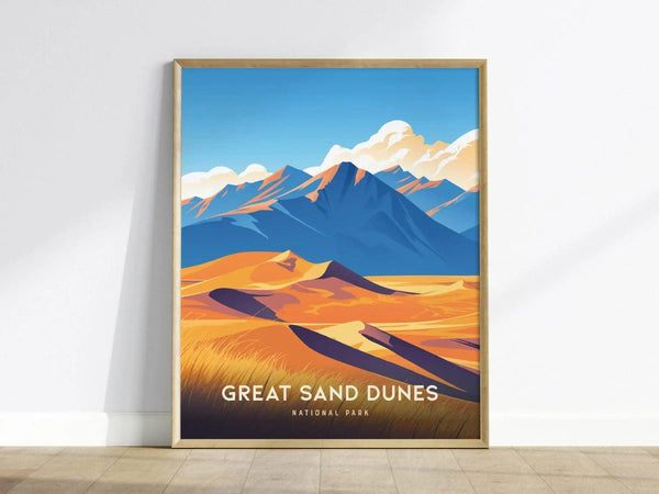 A framed poster of Great Sand Dunes National Park features vibrant golden dunes and blue mountains under a clear sky, leaning against a white wall on a wooden floor.