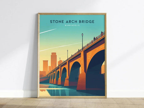 Framed poster of the Stone Arch Bridge in Minneapolis, featuring a stylized sunset skyline and reflections in the Mississippi River, leaning against a white wall on a wooden floor.