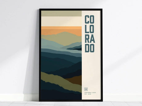 Framed minimalist poster featuring abstract layers of mountain ranges in blue and orange hues, with the word COLORADO vertically on the right. The poster sits on a white floor.