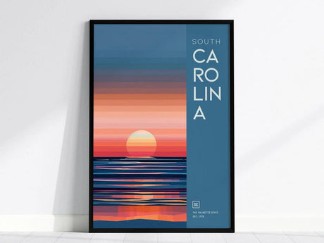 A framed poster featuring an abstract sunrise with warm hues over water, labeled South Carolina on the right, set against a light-colored wall.