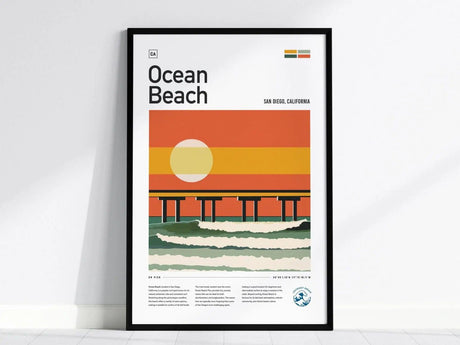 Framed poster featuring a stylized illustration of Ocean Beach in San Diego, showing a pier at sunset with vibrant orange and yellow hues, placed against a plain white wall.
