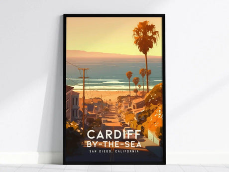 Framed poster depicting Cardiff-by-the-Sea, California, with a coastal view featuring palm trees, houses, and the ocean under a warm, golden sunset. Text reads CARDIFF BY-THE-SEA SAN DIEGO, CALIFORNIA.