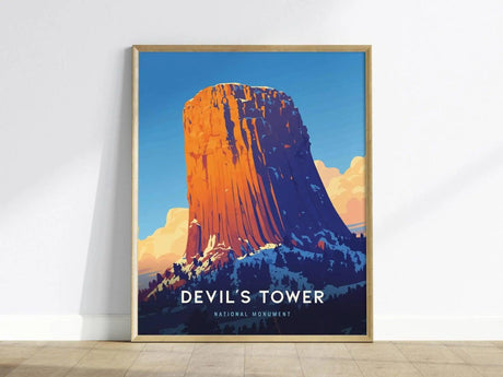 A framed poster of Devils Tower, featuring a vibrant illustration of the geological formation against a blue sky with clouds. Text reads Devils Tower National Monument.