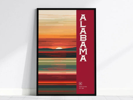 A framed minimalist poster featuring abstract horizontal lines in warm hues with the word Alabama in bold letters on the right. The sun sets behind a stylized landscape.