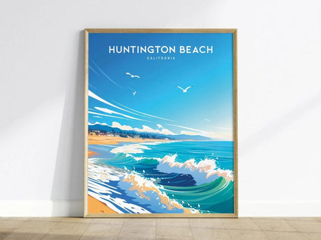 A framed Huntington Beach poster features a stylized ocean scene with waves, sandy shoreline, and seagulls under a bright blue sky, leaning against a white wall.