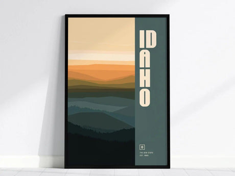 Framed Idaho poster featuring stylized mountains in shades of orange and teal with bold vertical IDAHO text on the right, displayed against a white wall.