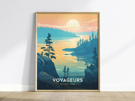 Framed poster of Voyageurs National Park depicting a serene lake scene at sunset, with silhouetted trees and gentle water reflections, leaning against a white wall on a wooden floor.