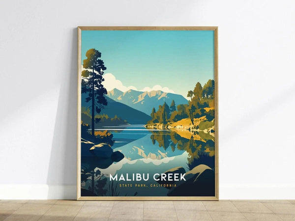 Framed poster of Malibu Creek State Park showing a serene lake with reflections of mountains and trees under a clear sky, resting against a white wall.