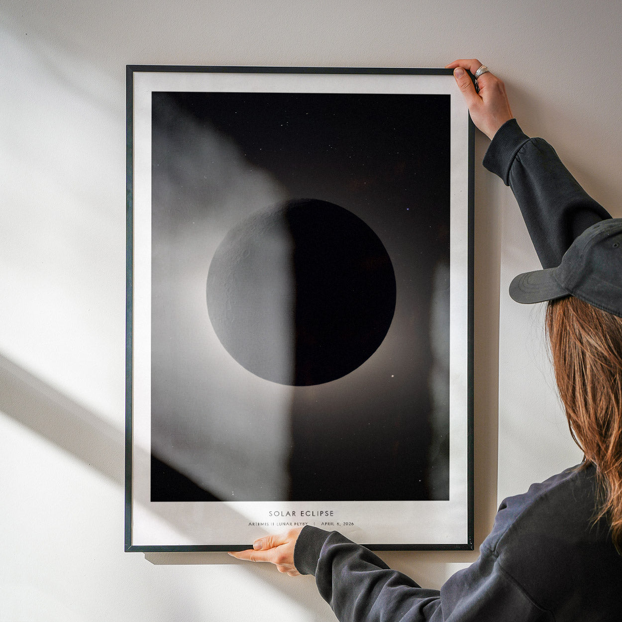 A person holds a framed art print of a solar eclipse against a wall. The print features a dark circular shape representing the moon, surrounded by a gradient of black and gray tones, creating a dramatic visual effect.