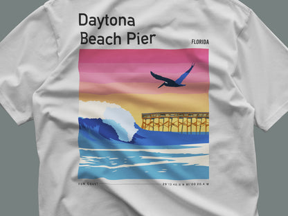 A white t-shirt features a vibrant graphic of the Daytona Beach Pier against a colorful sunset backdrop with shades of pink, orange, and blue. A bird soars above the waves, capturing the essence of Floridas coastal vibe.