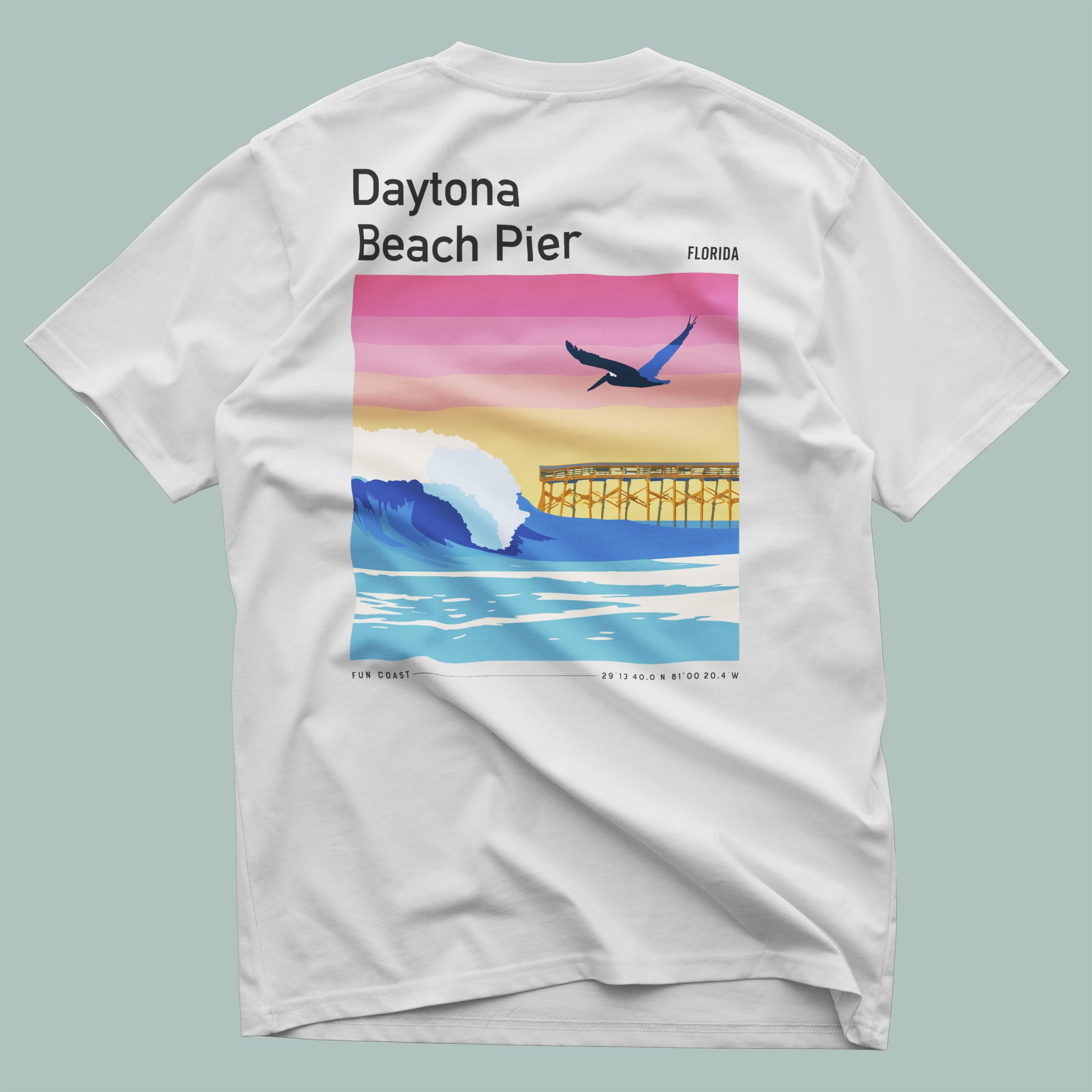 A white t-shirt features a vibrant graphic on the back showcasing the Daytona Beach Pier. The design includes colorful waves, a silhouette of the pier, and a bird soaring above, capturing the essence of a sunny beach atmosphere.