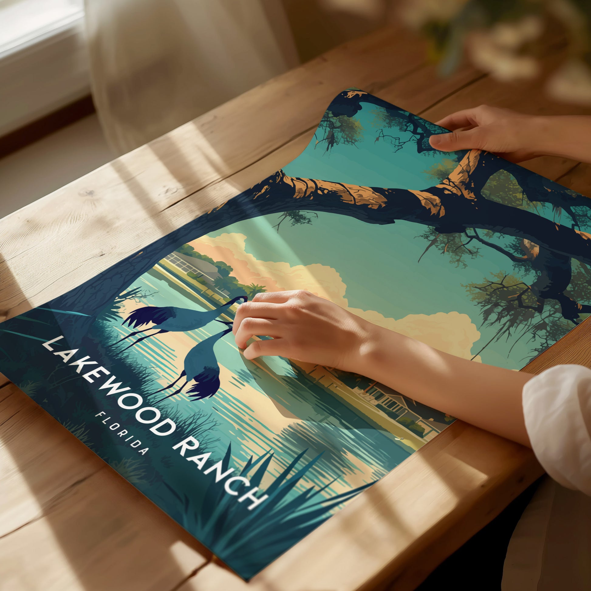 A person is holding a Lakewood Ranch poster depicting a scenic lakeside view with trees and birds, on a wooden table. Sunlight filters through a nearby window.