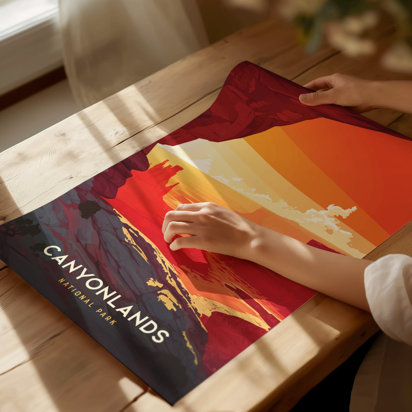 A person is holding a vibrant, colorful poster of Canyonlands National Park on a wooden table, featuring abstract desert and canyon landscapes. Sunlight streams through a nearby window.