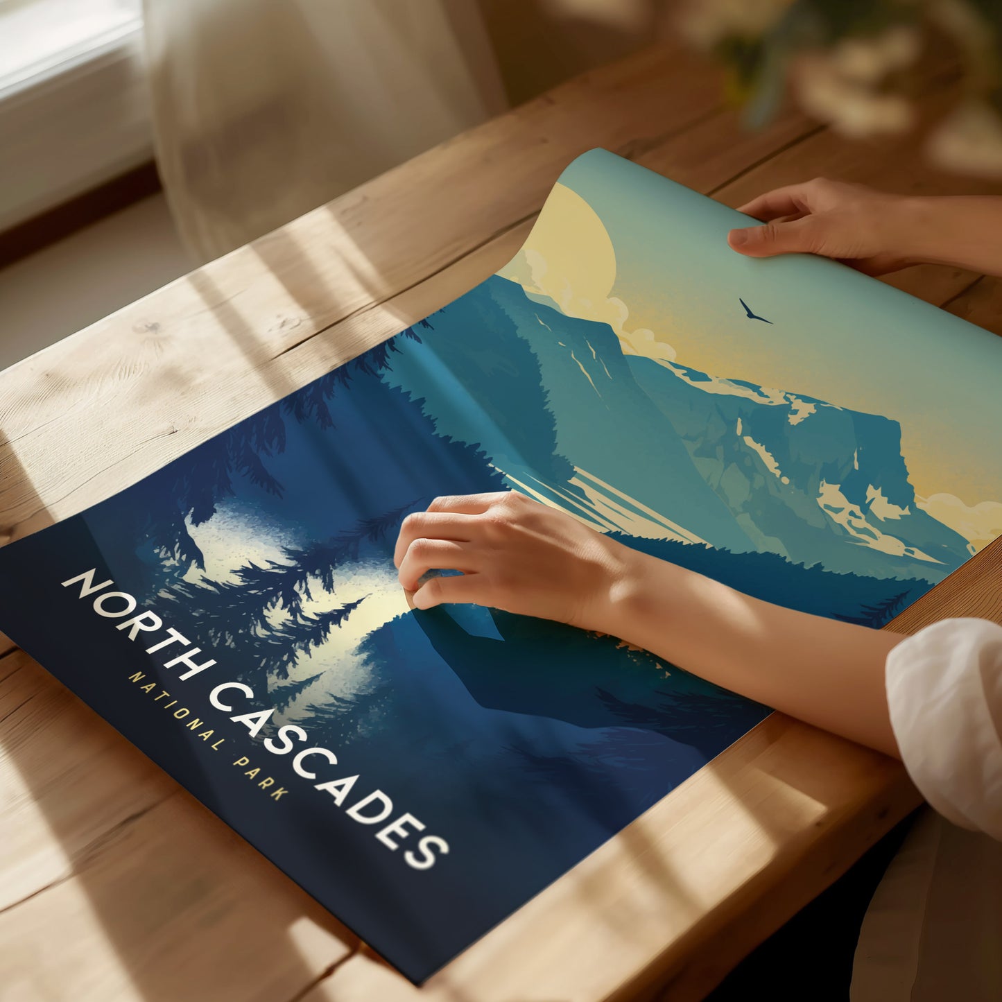 A person unrolls a North Cascades National Park poster on a wooden table, featuring stylized mountains, trees, and a bird in flight under a blue sky.