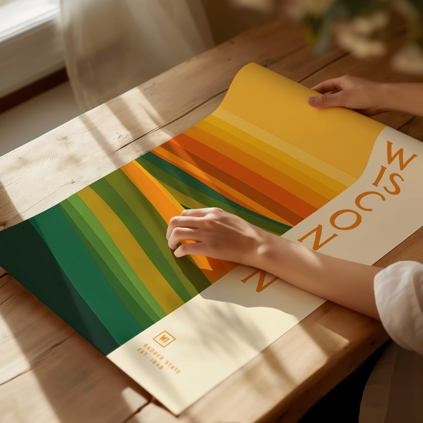 A person unrolls a colorful poster with abstract green and gold stripes on a wooden table. The poster says Wisconsin.