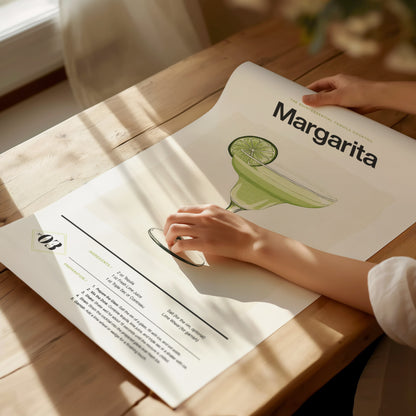 A person rolls a Margarita cocktail poster on a wooden table, featuring an illustration of a green margarita glass with a lime slice. Sunlight casts soft shadows across the scene.
