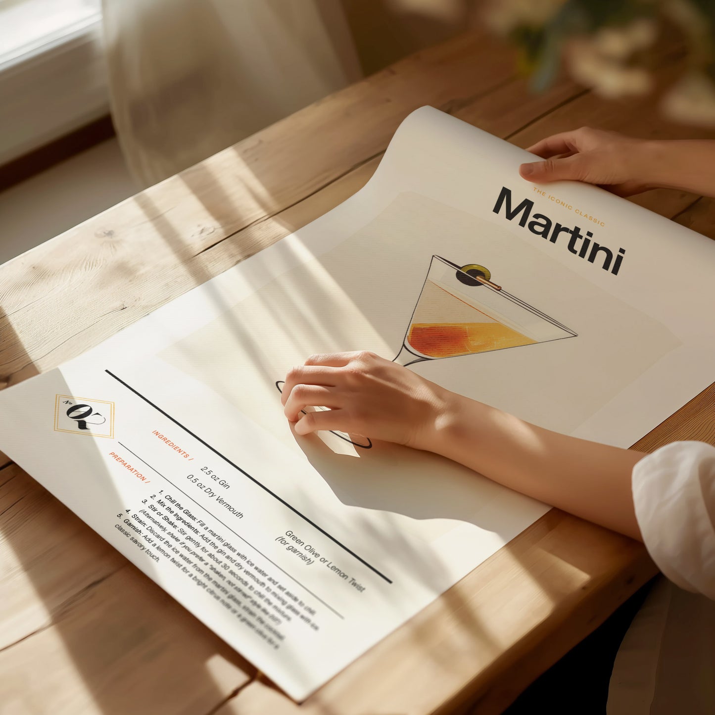 A person unrolls a Martini cocktail poster on a wooden table. The design features an illustration of a martini glass with a recipe and ingredients list.