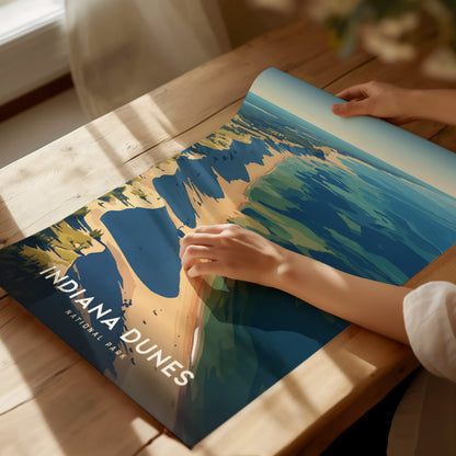A person unrolls an Indiana Dunes National Park poster on a wooden table, showcasing colorful illustrations of dunes.