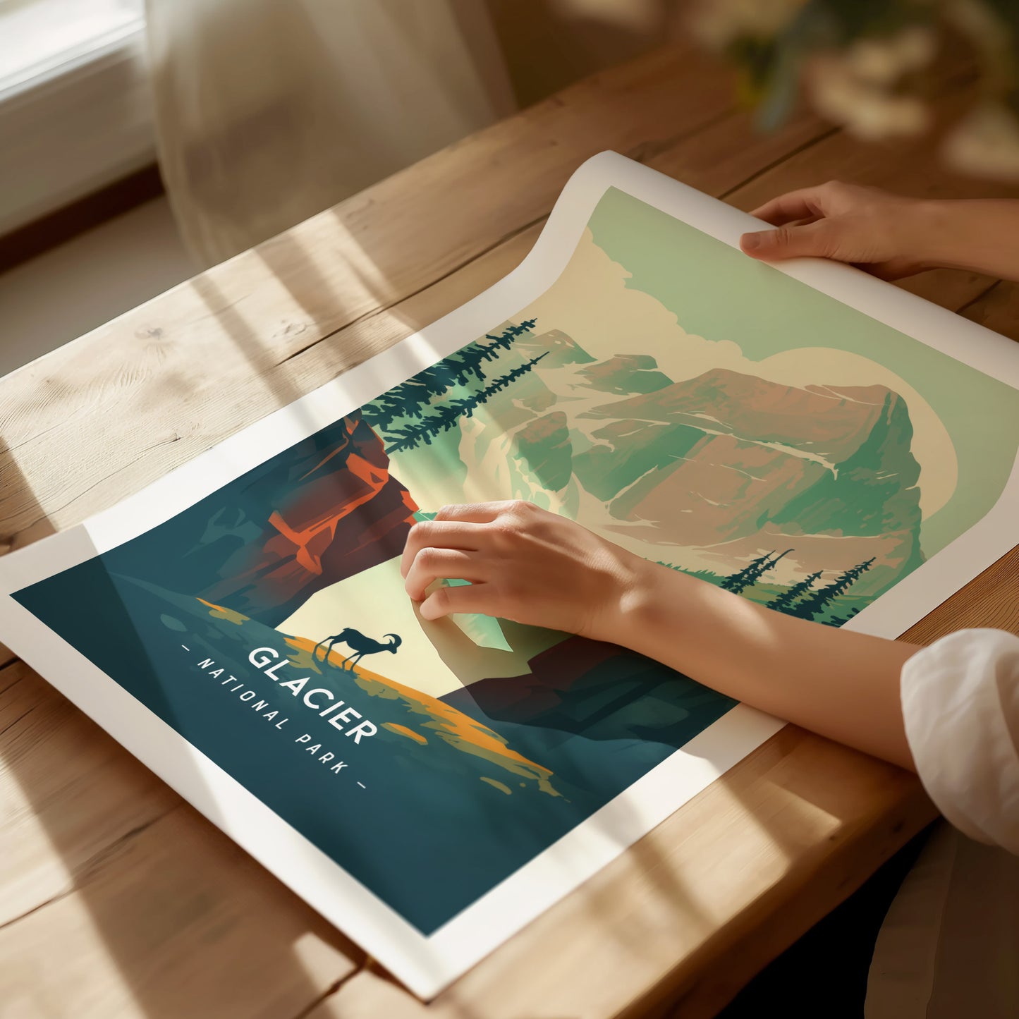 A person unrolls a colorful Glacier National Park poster on a wooden table, featuring a minimalist landscape with mountains, trees, and a bighorn sheep silhouette.